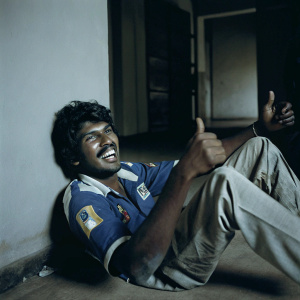 Akila Dananjaya, Sri Lankan cricketer, poses for a candid photo. Grinning widely, thumbs up gesture. Sporty attire, blue jersey with logos and patches.
