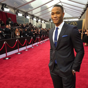 Trai Byers, dressed in a sharp charcoal suit with a white shirt and blue tie, standing at the Oscars nominee press conference. Red carpet and paparazzi in the background.
