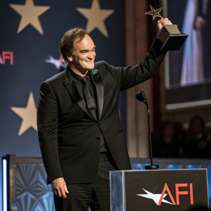 Quentin Tarantino in a sleek black tuxedo, accepting an award at the AFI Life Achievement Award. He beams as he stands on the stage surrounded by accolades and banners.