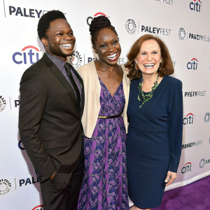 At PaleyFest, Ed Week, Xosha Roquemore, and Beth Grant, stars of 'The Mindy Project', beam with smiles on a purple carpet. Formal wear and cheerful expressions. White backdrop with text and logos.