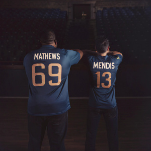 Cricket stars Angelo Mathews and Kusal Mendis posing at an indoor sports hall. They wear blue jerseys with numbers. Mathews sports number 69, while Mendis wears number 13. No audience visible.