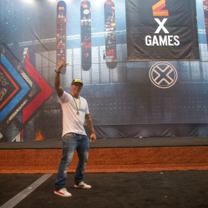 Ryan Sheckler, professional skateboarder, at X Games. Wearing a white graphic t-shirt, jeans, and sneakers. Vibrant backdrop featuring skateboard and X Games logos.