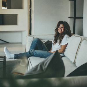 Tiffani Thiessen as Kelly Kapowski. Modern photo. Sitting on a futuristic sofa in a minimalist room. Wearing a casual white t-shirt and jeans, relaxed pose, bright smile.