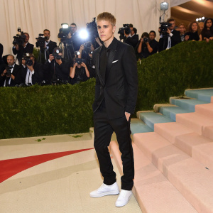 Justin Bieber at the Met Gala, dressed in a black suit with white sneakers. Crowd in the background, bright lights and paparazzi