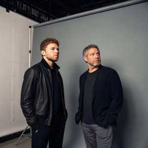Ryan Phillippe and Steven Silver in a stark studio setting. Both dressed in dark-toned attire. Simple gray backdrop