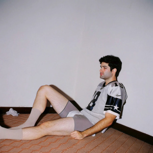 Jarrett Stidham in the locker room, wearing a casual jersey and shorts. Low-key lighting, teammate in background