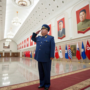 Kim Jong Un, dressed in a blue military uniform, stands in a vast hall decorated with national flags and portraits of leaders. He salutes the crowd.