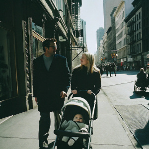 Mark Consuelos and Kelly Ripa strolling on a sunny street, baby in a stroller, vibrant urban backdrop.