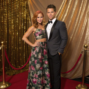 Amy Paffrath and Drew Seeley at a glitzy awards show. Amy in a trendy floral dress, Drew in a sharp tuxedo. They pose in front of a glamorous golden curtain backdrop.