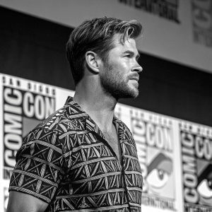 Chris Zylka at a Comic-Con panel. Black and white geometric print shirt. Toned physique, facial hair, serious demeanor. Colorful stage decorations backdrop.