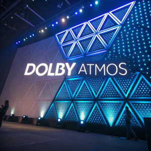 The Dolby Atmos logo in vivid detail. Modern geometric design with soft blue and dark tones. Illuminated against a dynamic backdrop of shifting light patterns and colors.