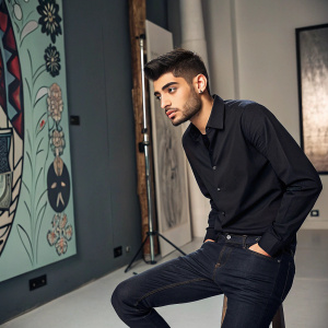 Zayn Malik, wearing a black shirt and dark jeans, posing alone in a studio with abstract artwork as the backdrop.