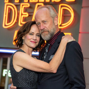 Sarah Clarke and Xander Berkeley embracing on The Walking Dead press tour. Both dressed in stylish outfits. Background features bright, neon signage
