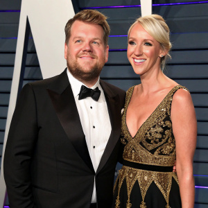 James Corden and Julia Carey, bearded actor and blonde actress, wearing elegant black and gold gowns. Posing happily at the Vanity Fair Oscar Party. The backdrop features a vibrant, geometric pattern with hints of blue and gray.