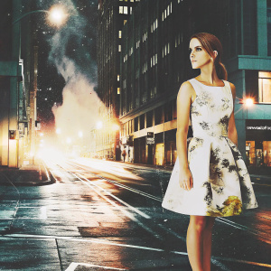 Emma Watson, in a stunning white dress with floral patterns, posing confidently on a movie set. The backdrop is a chaotic cityscape at night.