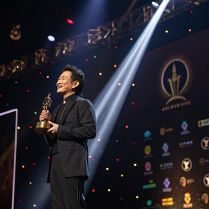 Alex Lim Hung-Fung, Asian actor, receiving award. Glowing spotlight, dramatic shadows, stage full of logos and decorations.