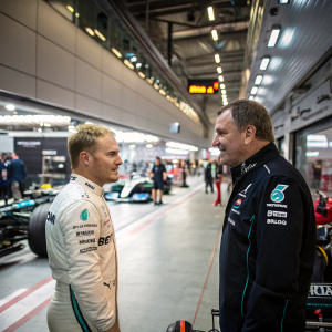 Valtteri Bottas and Nigel Mansell chatting at the Formula One team garage. Industrial machinery and mechanics bustling around in the background.