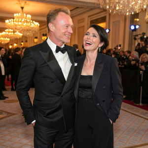 Gary Kemp and Sadie Frost at a chic gala. Both dressed formally. Gary in a black tuxedo, Sadie in a stylish black blazer and matching trousers. They share a joyful moment as they pose for photographers amidst a crowd of guests inside a grand ballroom.
