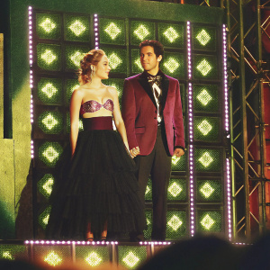 At the vibrant Nickelodeon Kids' Choice Awards Mexico 2019, a young Caucasian woman in a stunning strapless top with ruffled details and a tiered black skirt stands beside a man with South Asian features, dressed in a maroon blazer and formal attire. The stage backdrop is filled with bright Nickelodeon logos.