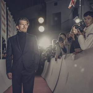 Ethan Hawke, dressed in a black turtleneck and tailored navy suit, amidst a vibrant crowd at a movie premiere.