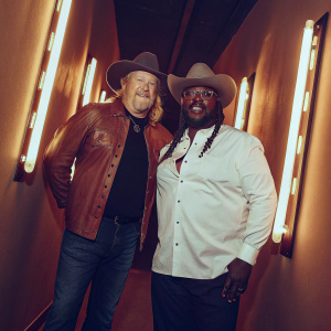 Trace Adkins and T-Pain, both in cowboy hats, posing for a candid backstage photo at the Country Music Awards. Neon lights in the background