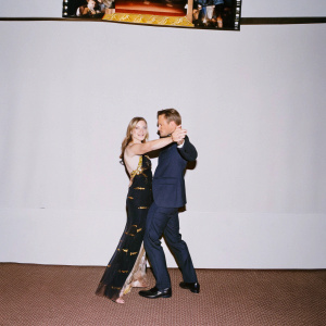 Anna Torv and Mark Valley at a sci-fi convention. Anna, in a flowing black gown with gold accents, and Mark in a sharp navy suit. Standing before a vibrant Star Trek banner. The backdrop teems with flashing lights and excited fans.
