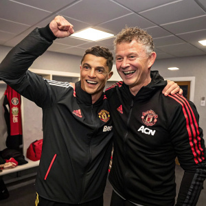 Cristiano Ronaldo and manager Ole Gunnar Solskjær celebrating victory in post-match interview room. Ronaldo with tousled hair, smiling broadly. Both dressed in training gear with team emblem.