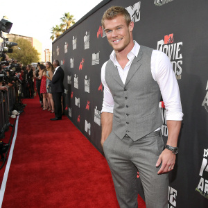 Alan Ritchson, actor, on the MTV Movie Awards red carpet. Grey vest, white shirt, grey pants. Black backdrop with MTV logos.
