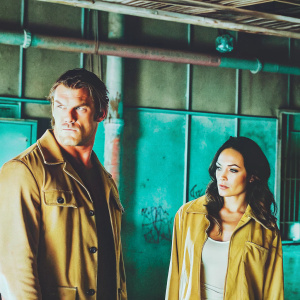 Alan Ritchson and Christina Ochoa on set of Blood Drive. Dark, gritty warehouse interior with industrial pipes and graffiti. Both actors have serious expressions.