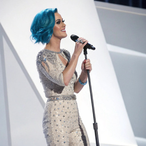 Katy Perry at the Grammy Awards, blue hair, standing alone on stage with a microphone in hand, white background
