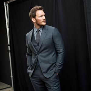 Chris Pratt in promotional photoshoot for new movie. Wears a stylish charcoal grey suit, solid black backdrop, confident expression