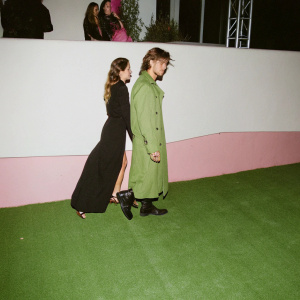 Paris Jackson and Jordan Barrett at a casual street style event. Paris in a black dress, Jordan in a greenish-brown trench coat-like garment. Various stylish attendees in the background.