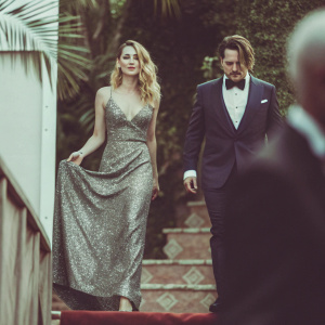 Amber Heard and Johnny Depp at a black tie gala. She is in a floor-length, silver sequined dress with a plunging neckline and he is in a tailored navy tuxedo. Luxurious gold backdrop