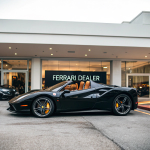 Black Ferrari 488 Spider parked outside a luxury car dealership. Dealer's nameplate visible, sign reads 'Ferrari Dealer'. Shiny black exterior, open-top design. Brown leather interior, sleek lines
