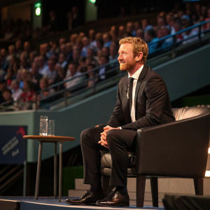 Paul Collingwood, cricket player, in a formal black suit. Sitting on stage during a Q&A session at a sports conference.