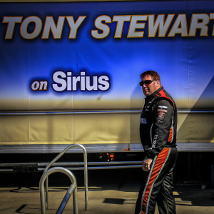 Tony Stewart, in racing gear with sponsor patches, stands at a NASCAR event. A fiery backdrop with his name and 'on sirius' emblazoned.