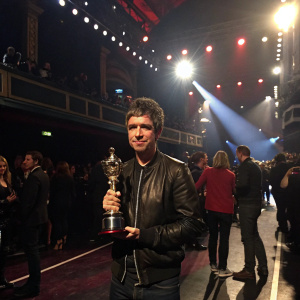 Noel Gallagher, rock star, holding trophy. Award ceremony at O2 Academy, Brixton. Crowded indoor event, bright stage lights.
