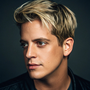 Close-up of Milo Yiannopoulos, youthful Caucasian male with short blonde hair, neutral expression. Shot in studio with controlled lighting. Dark blurred background highlighting facial features.