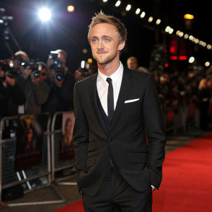 Tom Felton, dressed in black suit and tie, at the premiere event. Red carpet and flashing cameras in the background.