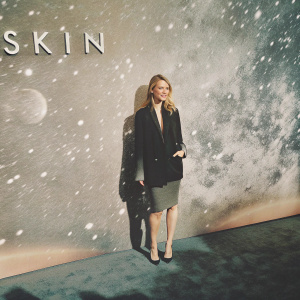 Alona Tal at the film 'Skin' premiere. Wearing a black blazer with matching pencil skirt. Standing in front of a backdrop featuring the film's promotional logos and a starry night sky