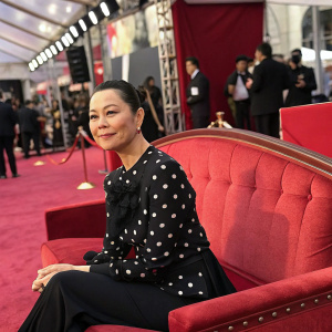 China Chow, sitting elegantly on a vintage red sofa, wears a black blouse with white polka dots. She exudes poise and charm amidst the bustling backstage area at a glamorous event.
