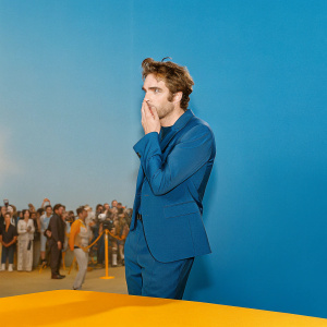 Robert Pattinson in a candid moment at the Venice Film Festival. Dressed in a navy blue suit, looking thoughtful. Crowded and bustling event backdrop.