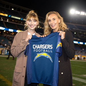 Imogen Poots and Toni Collette posing with a Los Angeles Chargers football jersey at a promotional event. The jersey features the Chargers logo and the text 'Chargers Football' in a sporty font. The event is set in a stadium.