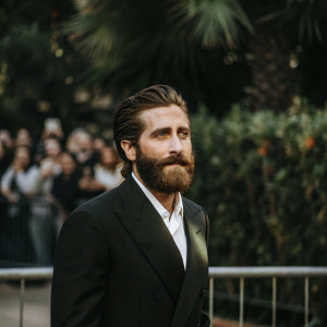 Jake Gyllenhaal, sporting a full beard, dressed in a black suit and white shirt, stands in front of a blurry crowd at a film festival.
