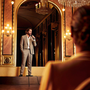 Jake Gyllenhaal at a gala event. Speaking into a microphone. Lavish room with chandeliers and ornate decorations.
