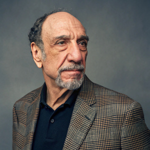 Professional headshot of F. Murray Abraham, a distinguished actor. Gray gradient background. Wears a plaid blazer with dark shirt underneath. Mustache prominent. Upper body shot, neutral studio setting.