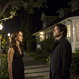 Jennifer Love Hewitt and David Conrad from 'Ghost Whisperer'. Jennifer, wearing a black dress, and David, in a dark jacket, standing outside a house at night. The scene is illuminated by soft street lights and the glow from the house windows.