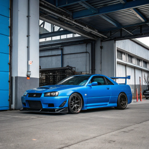 A picture of a Nissan Skyline R34 in vivid blue color, parked in an open garage. The car's sleek design and sporty features are prominently displayed. Background is grey with industrial details.
