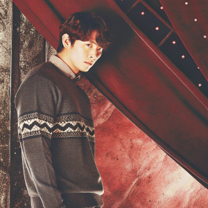 Korean actor Ji Sung, dressed in a dark gray sweater with zigzag pattern, posing solemnly against a dramatic black and red backdrop.