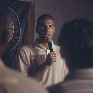 Zinedine Zidane addressing the media in a white polo shirt. The backdrop features a team logo.